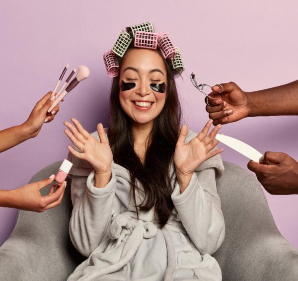 Cheerful brunette mixed race female takes care of face skin, reduces puffiness with eye patches, raises palms from happiness, wears hair curlers, comfortable bathrobe, enjoys beauty treatments